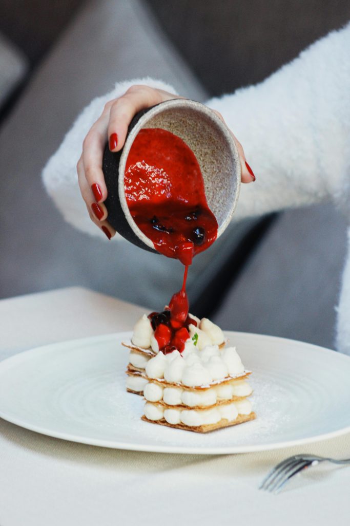 Close-up of a millefoglie dessert being topped with vibrant berry sauce, showcasing its layers.